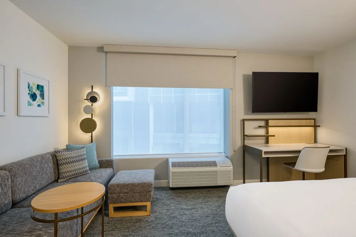 Hotel room with window, air conditioner, gray sofa, small wooden table, wall art, flat-screen TV, desk with chair.