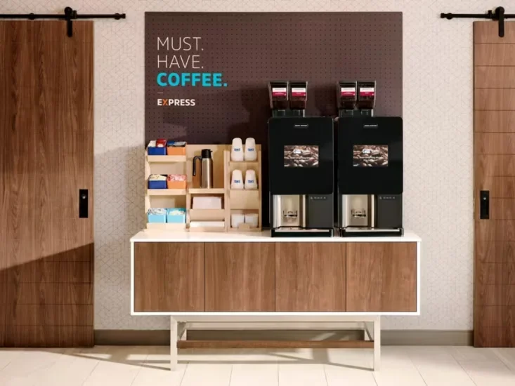 Coffee station with two coffee machines, cups, and supplies on a wooden counter, with a sign that says "Must Have Coffee" and sliding wooden barn doors on either side.