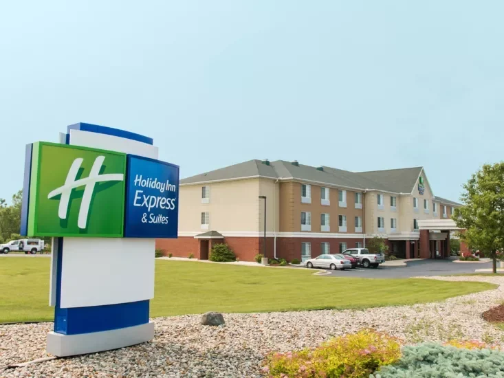 Exterior view of a Holiday Inn Express & Suites hotel with a large sign in the foreground and a beige hotel building in the background, surrounded by a well-kept lawn and parking lot.