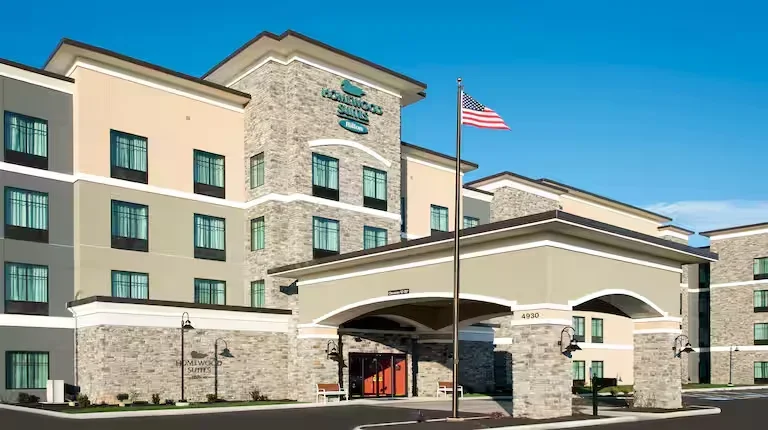 The exterior of a Sleep Inn & Suites hotel with an American flag flying in front, multi-story building with stone and beige siding, and a blue sky in the background.