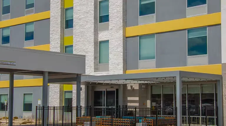 Modern apartment building with gray and yellow accents, large windows, and an outdoor seating area fenced with black metal, featuring benches and a canopy.