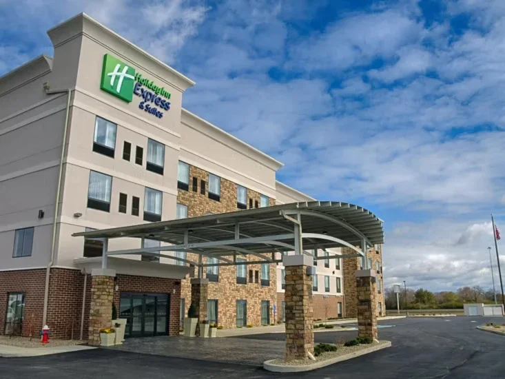 Exterior of a Holiday Inn Express & Suites hotel with a covered entrance, brick and beige siding, and a large sign on the building and flag in the background under blue sky with clouds.