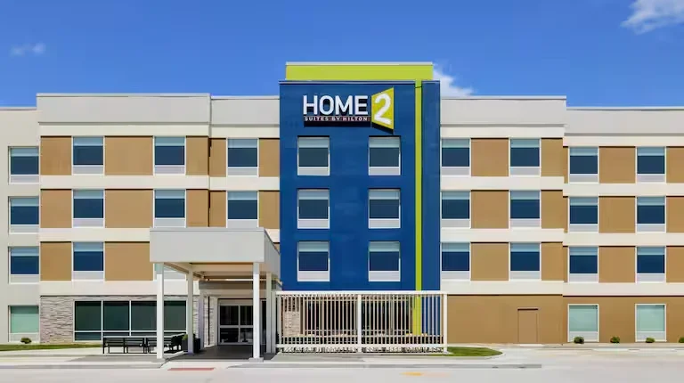 Exterior of a Home2 Suites by Hilton hotel, three-story building with beige and blue facade, main entrance with canopy, and a sign on top displaying the hotel name and logo.