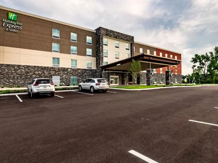 Exterior of a Holiday Inn Express hotel with a parking lot, featuring a covered entrance and parked cars, under a partly cloudy sky.