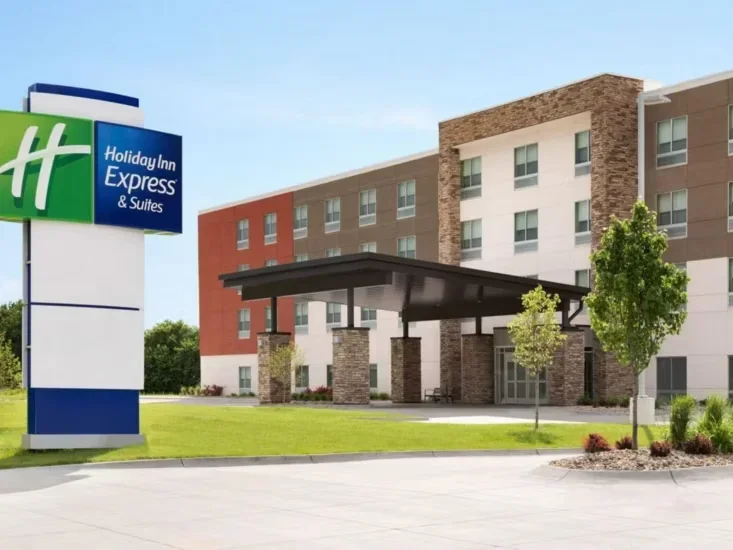 Exterior view of a Holiday Inn Express & Suites hotel with a large sign, entrance canopy, and landscaped surroundings during daytime.