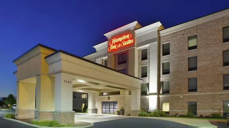 Exterior view of a Hampton Inn & Suites hotel at dusk, showing the entrance and the illuminated Hampton Inn & Suites sign on the building.