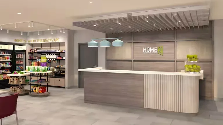 Reception area with a curved front desk, a sign reading "Home 2," shelves with grocery items, and a display of apples.