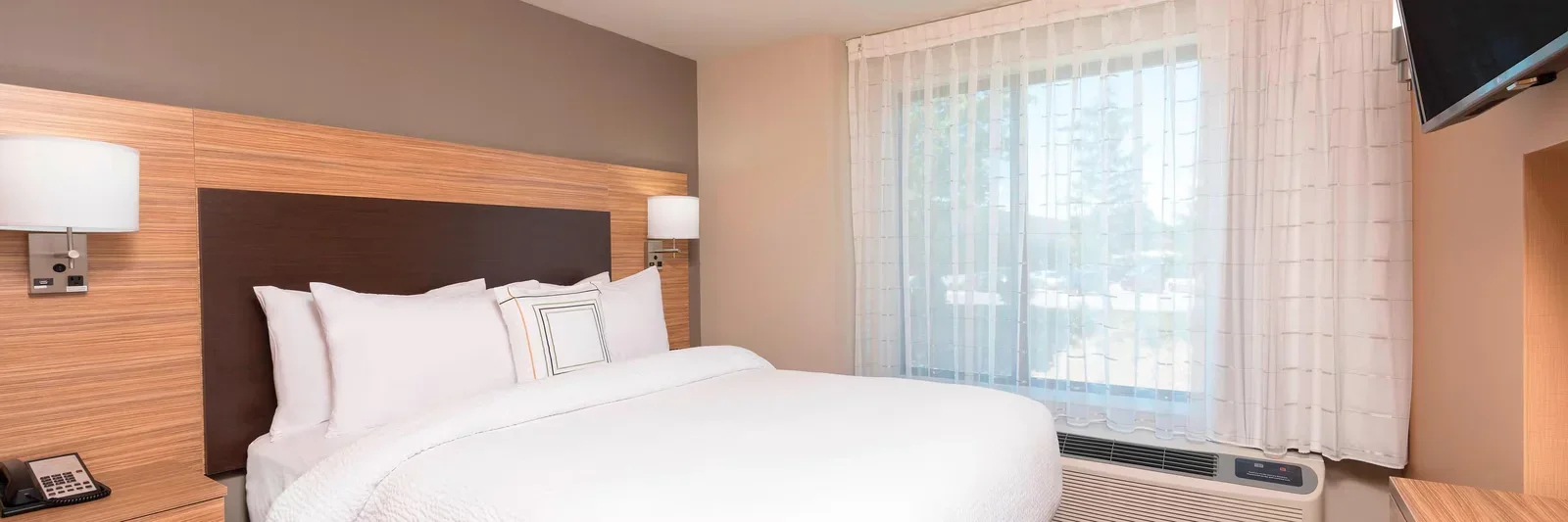 A hotel room with a bed, wooden headboard, white bedding, pillows, two wall-mounted lamps, a telephone, a window with sheer curtains, and a wall-mounted TV.