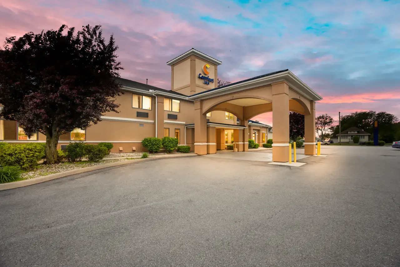 Exterior view of a Comfort Inn hotel at sunset with a covered driveway, landscaped bushes, a tree, and a colorful sky.