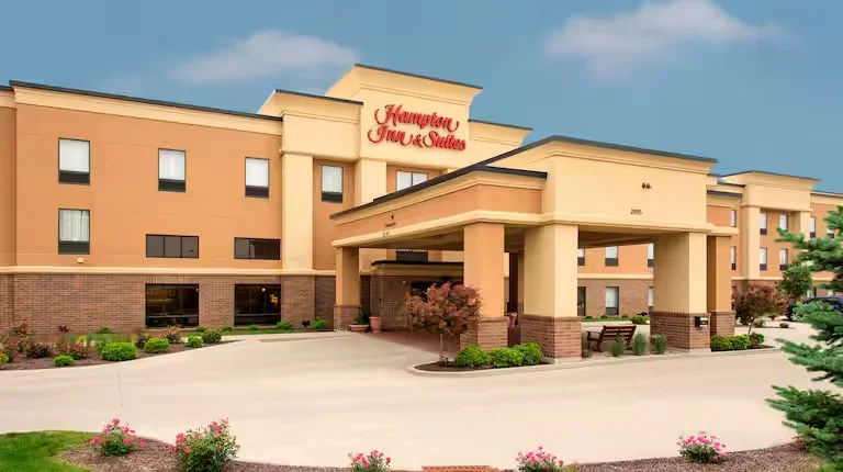 Hampton Inn & Suites hotel exterior with a beige facade, a covered entrance, and landscaping with bushes and flowers.