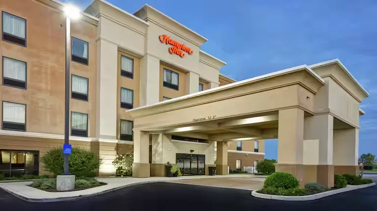 Hampton Inn hotel with a covered entrance and green landscaping under a blue sky.