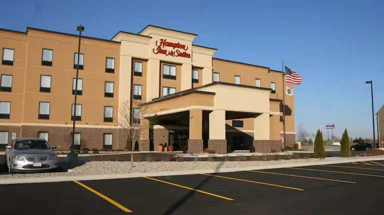 Exterior of Hampton Inn & Suites hotel with parking lot, American flag, and clear blue sky.