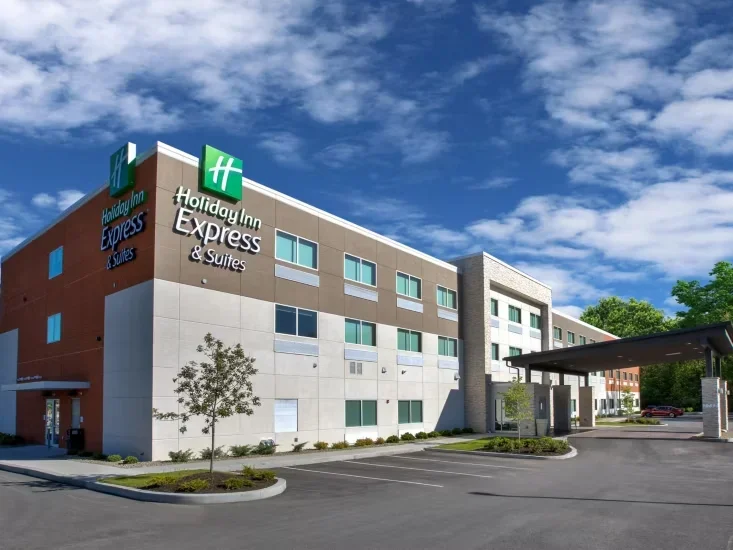 Exterior view of a Holiday Inn Express & Suites hotel, showing the building's modern architecture, parking lot, and a partly cloudy sky.