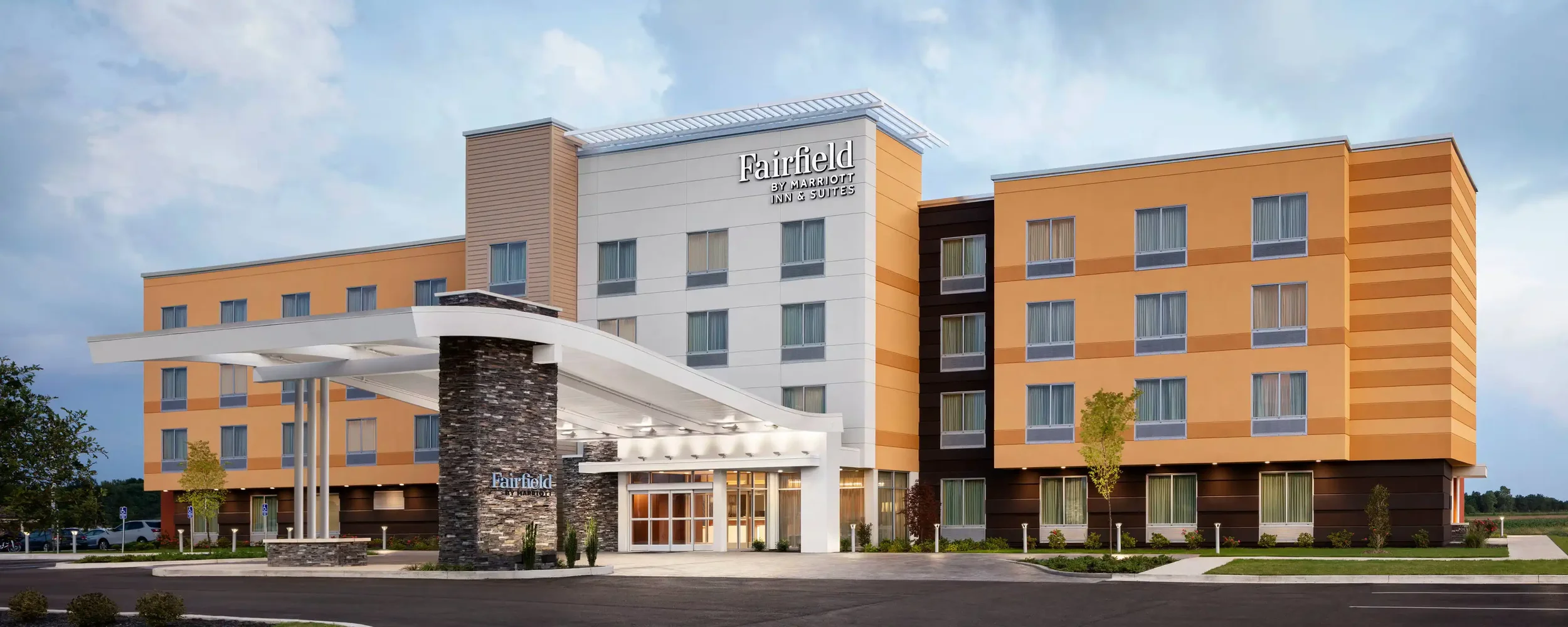 Exterior view of a Fairfield by Marriott Inn & Suites hotel with a modern architectural design, featuring a yellow and white facade, large front entrance with a glass door, and a landscaped area with small trees and shrubs.