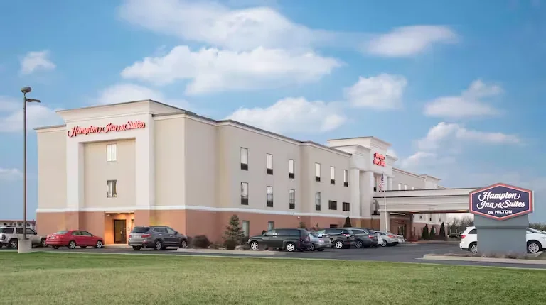 Exterior view of a Hampton Inn & Suites hotel with a parking lot in front, under a partly cloudy sky.