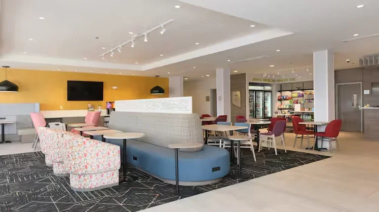 Modern lounge area with colorful chairs, sofas, and tables in a bright, open space with a yellow accent wall and a display with snacks.