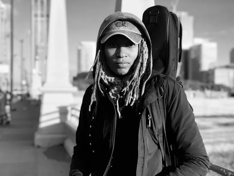 A woman with dreadlocks wearing a baseball cap and a black jacket, with a backpack on her shoulder, standing outdoors in an urban setting.