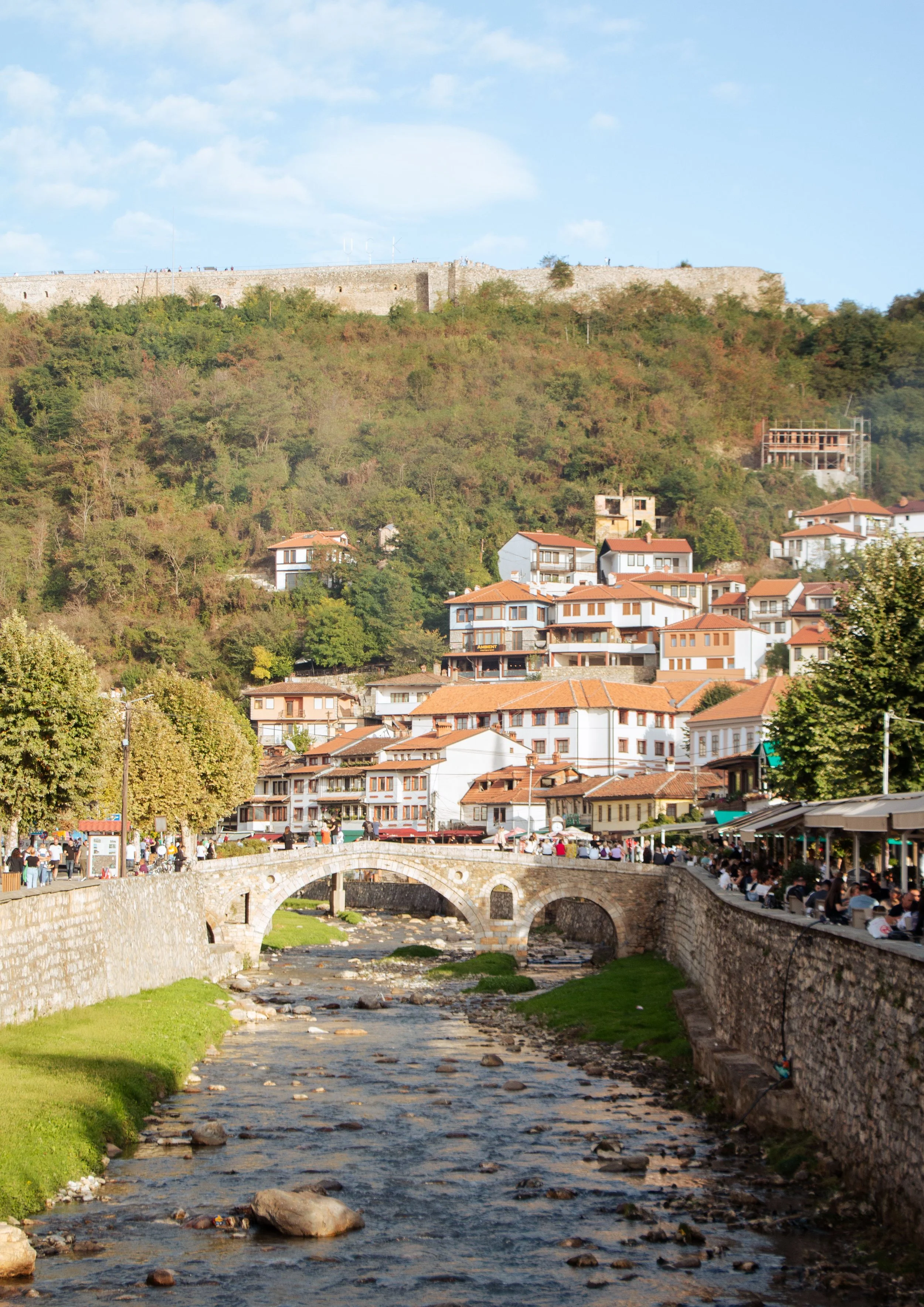 Blick auf eine Stadt mit Fluss, Brücke und Häuser im Vordergrund, mit einem Hügel und Burgmauer im Hintergrund. Viele Menschen sitzen an Bars am Fluss und es ist schönes Wetter.