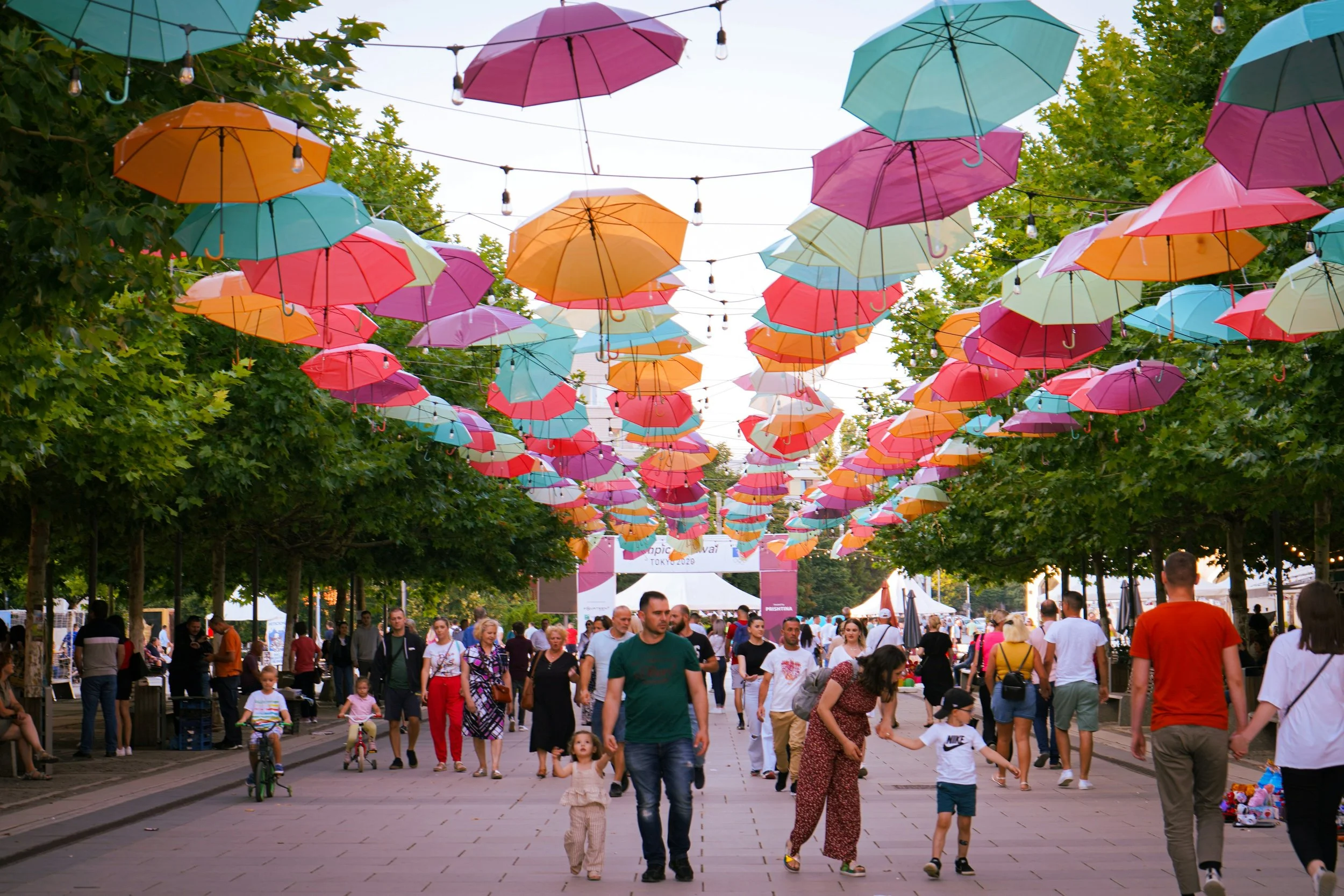 Bunte Regenschirme hängen über einer belebten Stadtstraße mit Passanten, Bäumen und Zelten im Hintergrund.