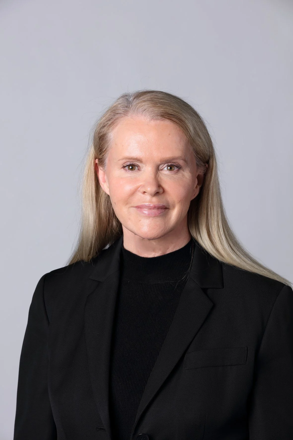 A portrait of a woman with long blonde hair wearing a black blazer and black turtleneck, smiling softly, against a plain gray background.
