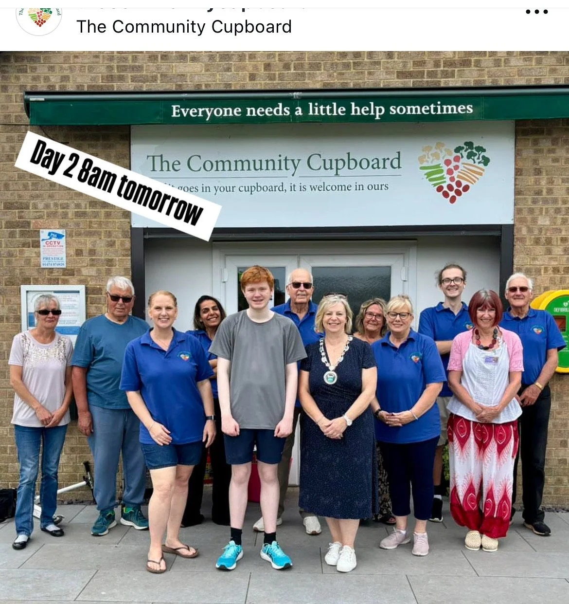 Group of people standing in front of a community cupboard with a sign that reads 'Everyone needs a little help sometimes'.