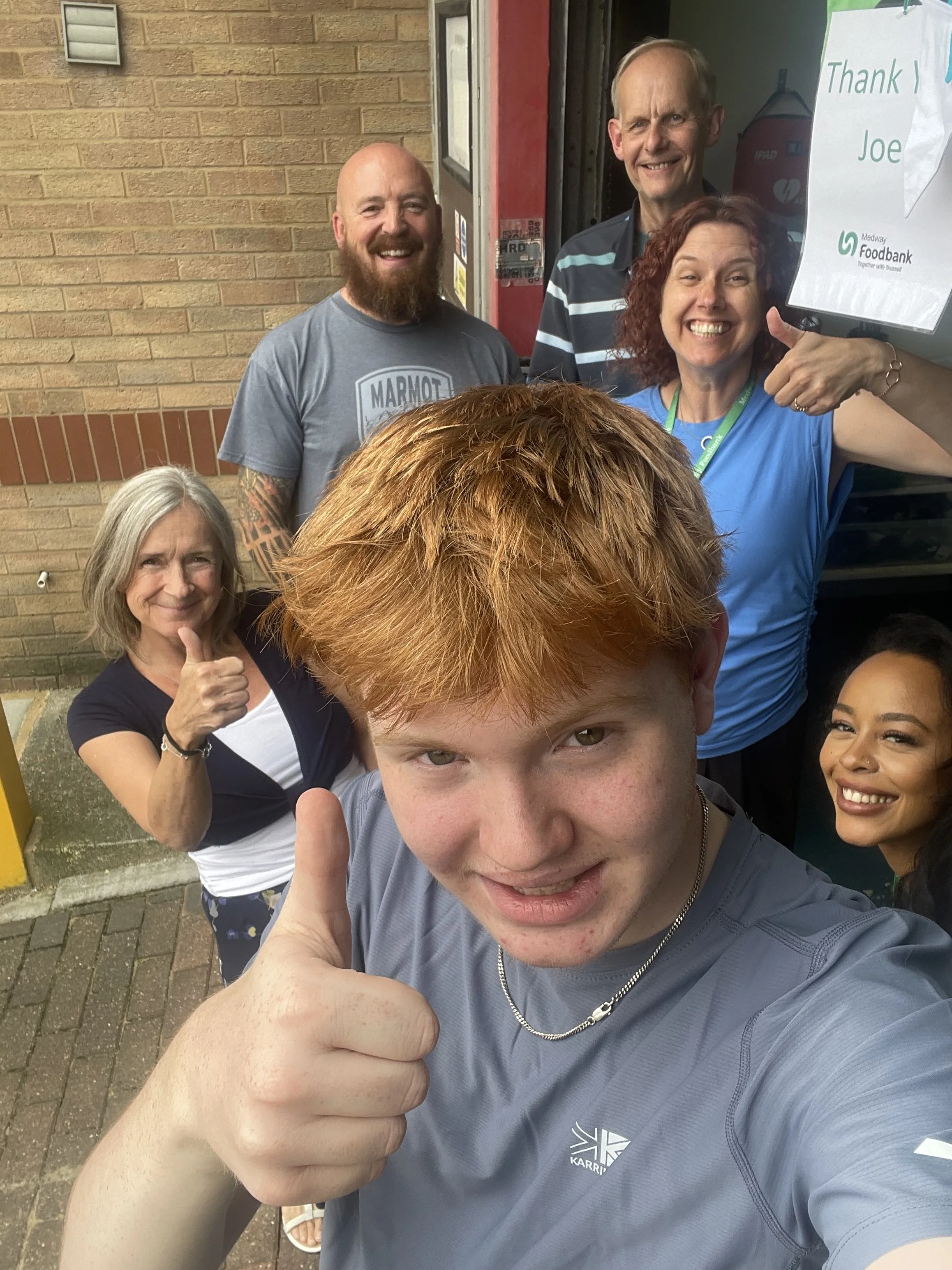Group of five people smiling and giving thumbs up outside near a brick wall. One person in the foreground with red hair, a person in the background with a gray beard, and three women, one with curly red hair, one with gray hair, and one with long dark hair.