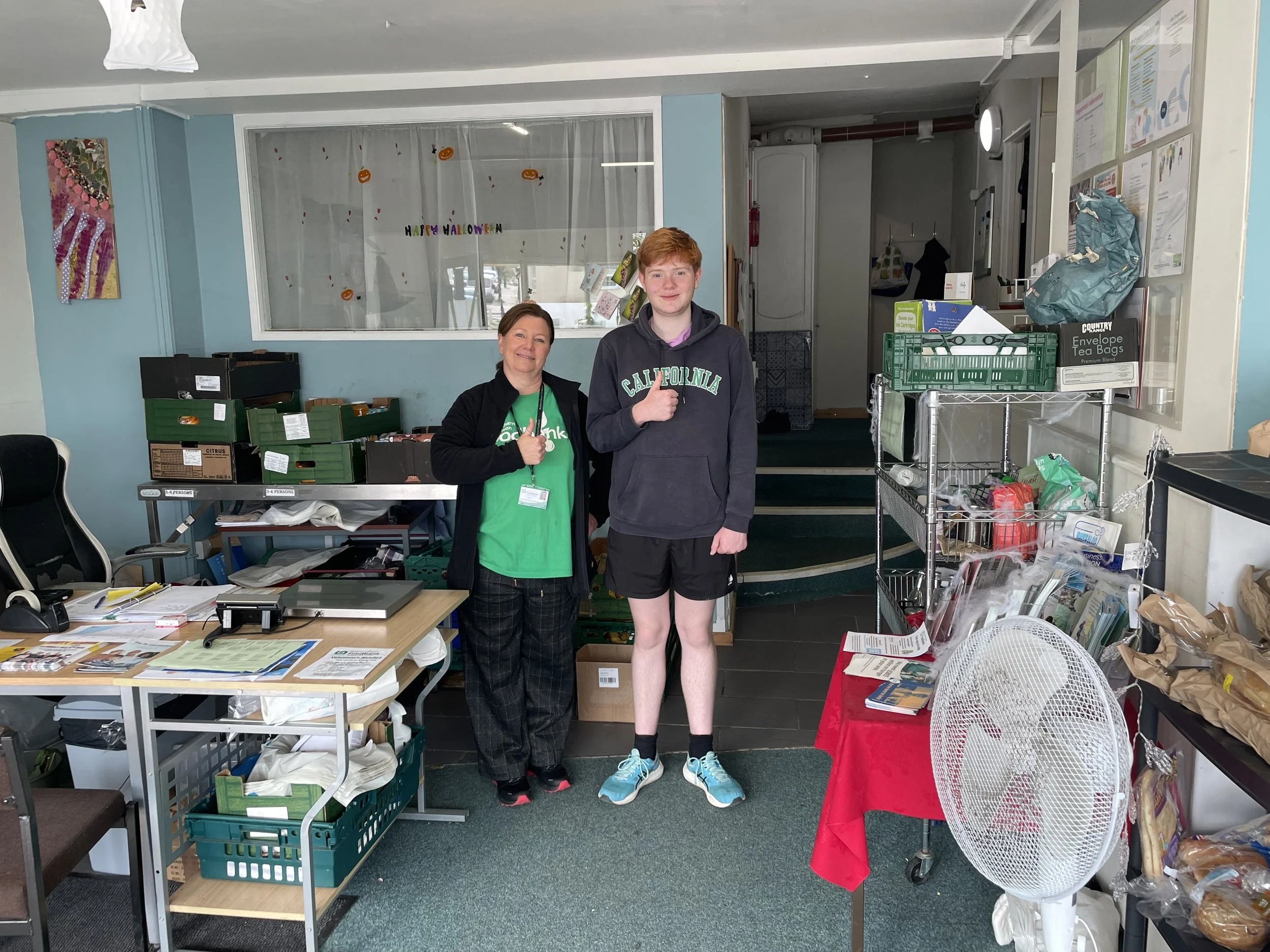 Two people, a woman and a young man, standing inside a room with various office supplies and decorations. The woman is giving a thumbs-up, smiling, and wearing a green shirt and dark jacket. The young man is also giving a thumbs-up, smiling, and dressed in a dark hoodie and shorts. Behind them, the wall has Halloween decorations, and there are tables and shelves filled with papers, boxes, and everyday items.