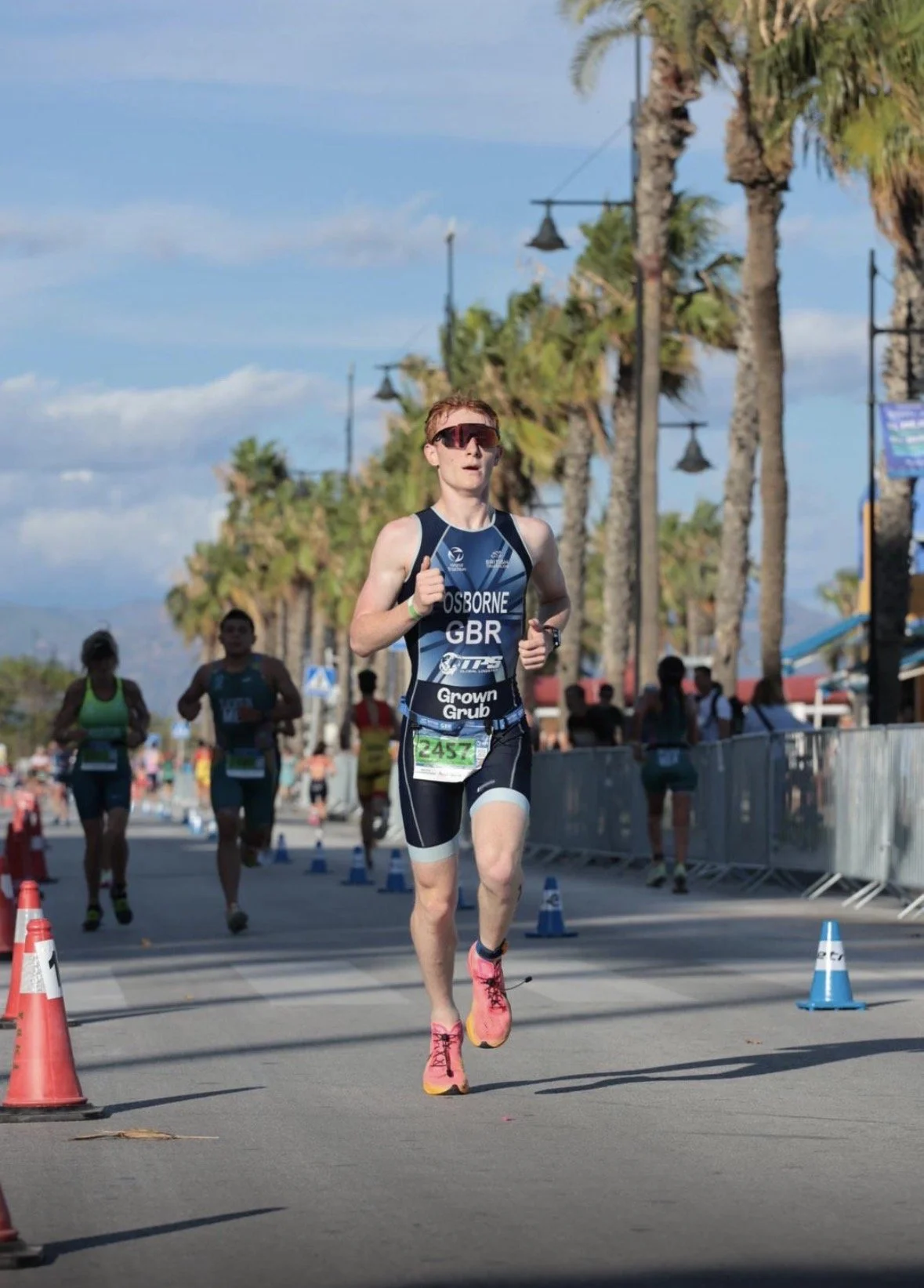 A male runner wearing sunglasses and a black and blue triathlon suit with the name 'OSBORNE' and 'GBR' on it, participating in a race outdoors on a sunny day, with palm trees and other runners visible in the background.
