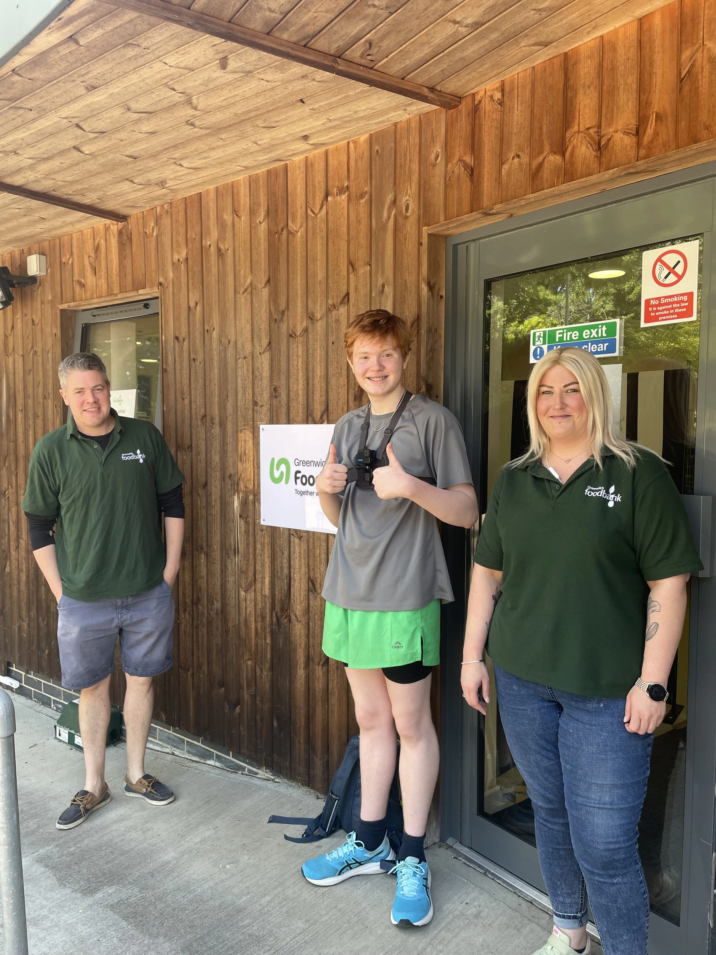 Three people standing outside a building with wooden siding, near a glass door with safety signs. Two are smiling and looking at the camera, one is giving a thumbs-up gesture.