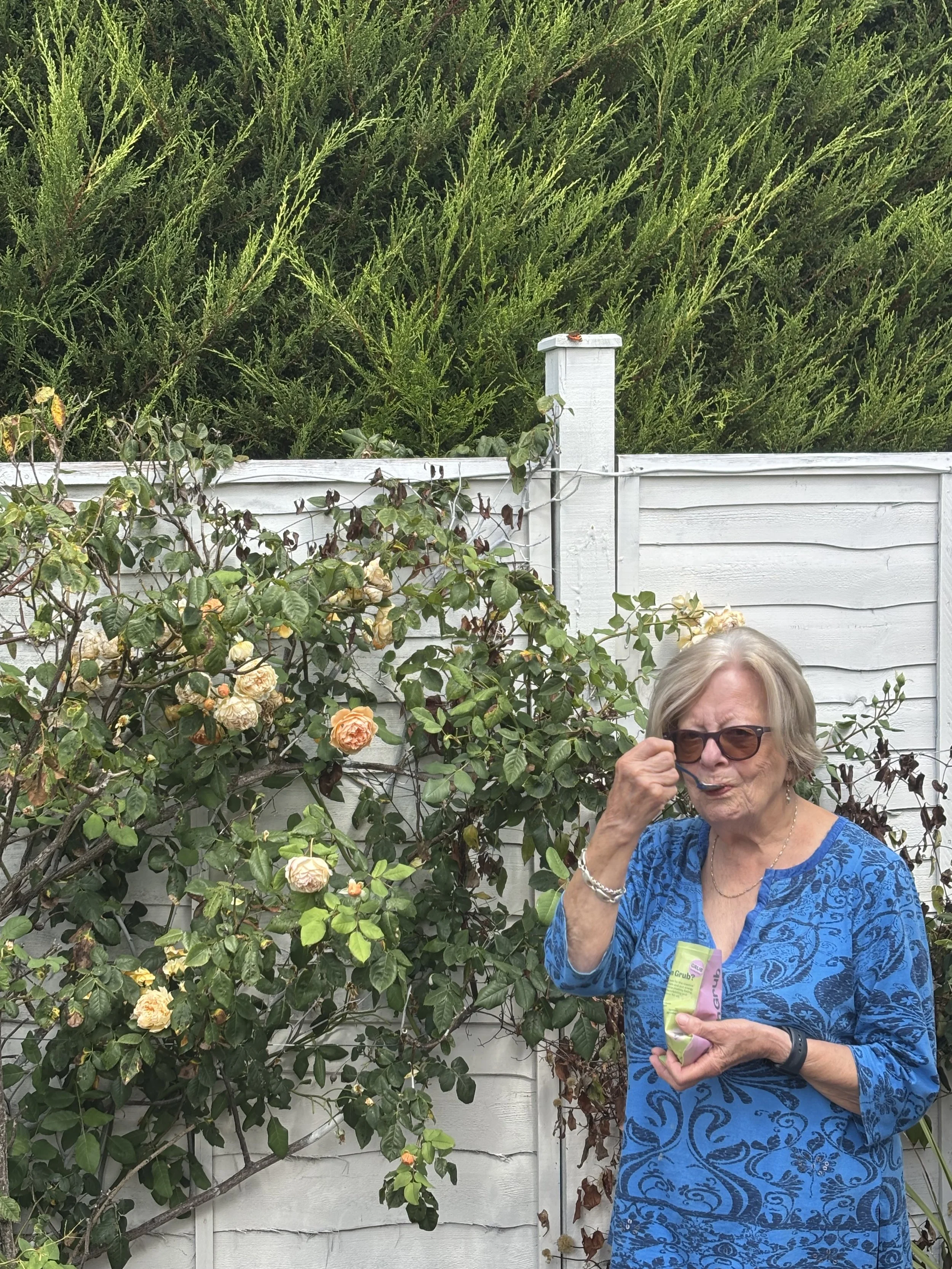 An elderly woman with short gray hair and glasses stands outdoors in front of a white wooden fence and green bushes, holding a snack bag and eating, wearing a blue patterned dress.