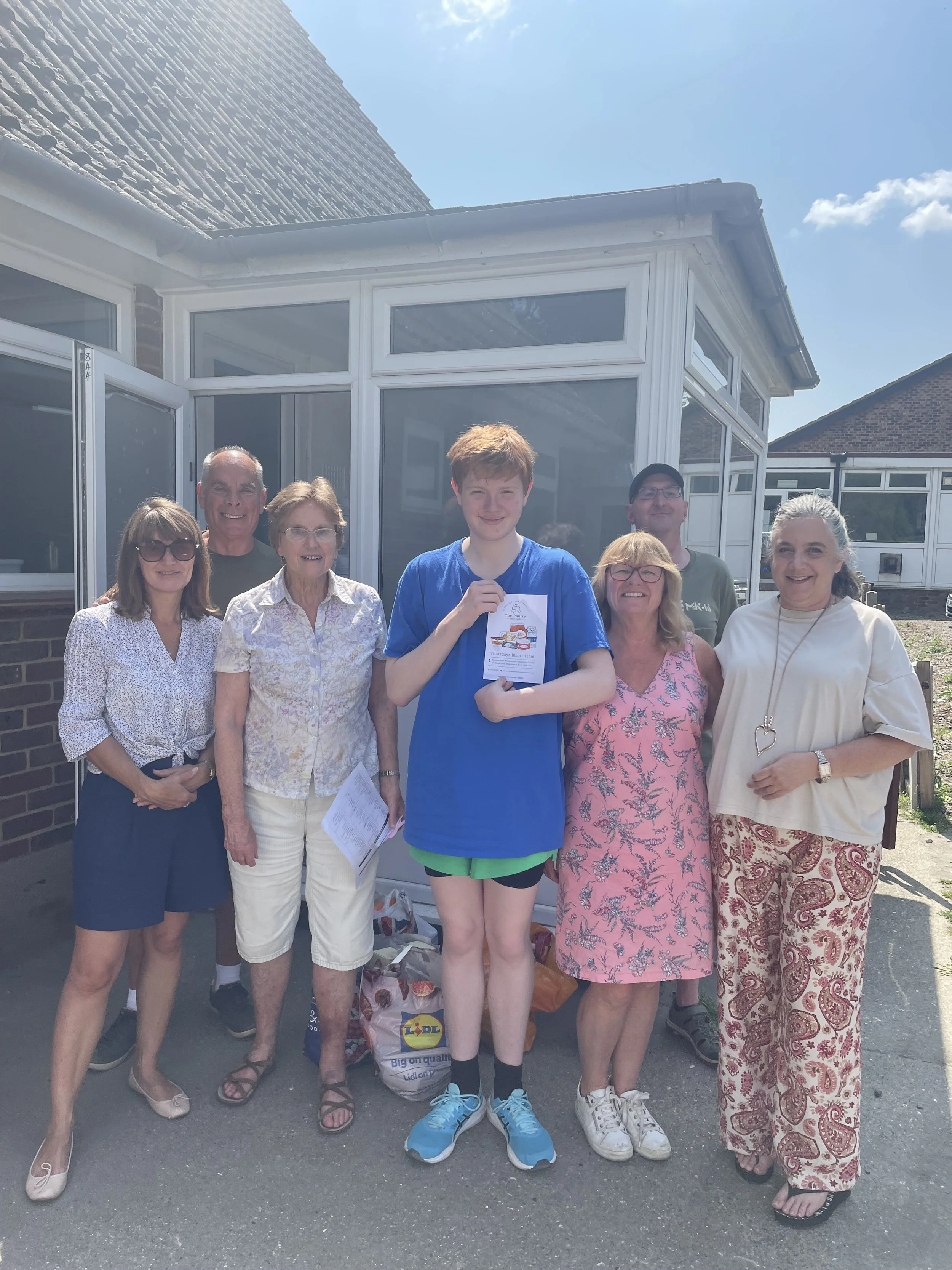 A group of eight people, including older adults and a young person, standing outside in front of a house with a conservatory. They are smiling and holding various items, on a sunny day with a clear blue sky.