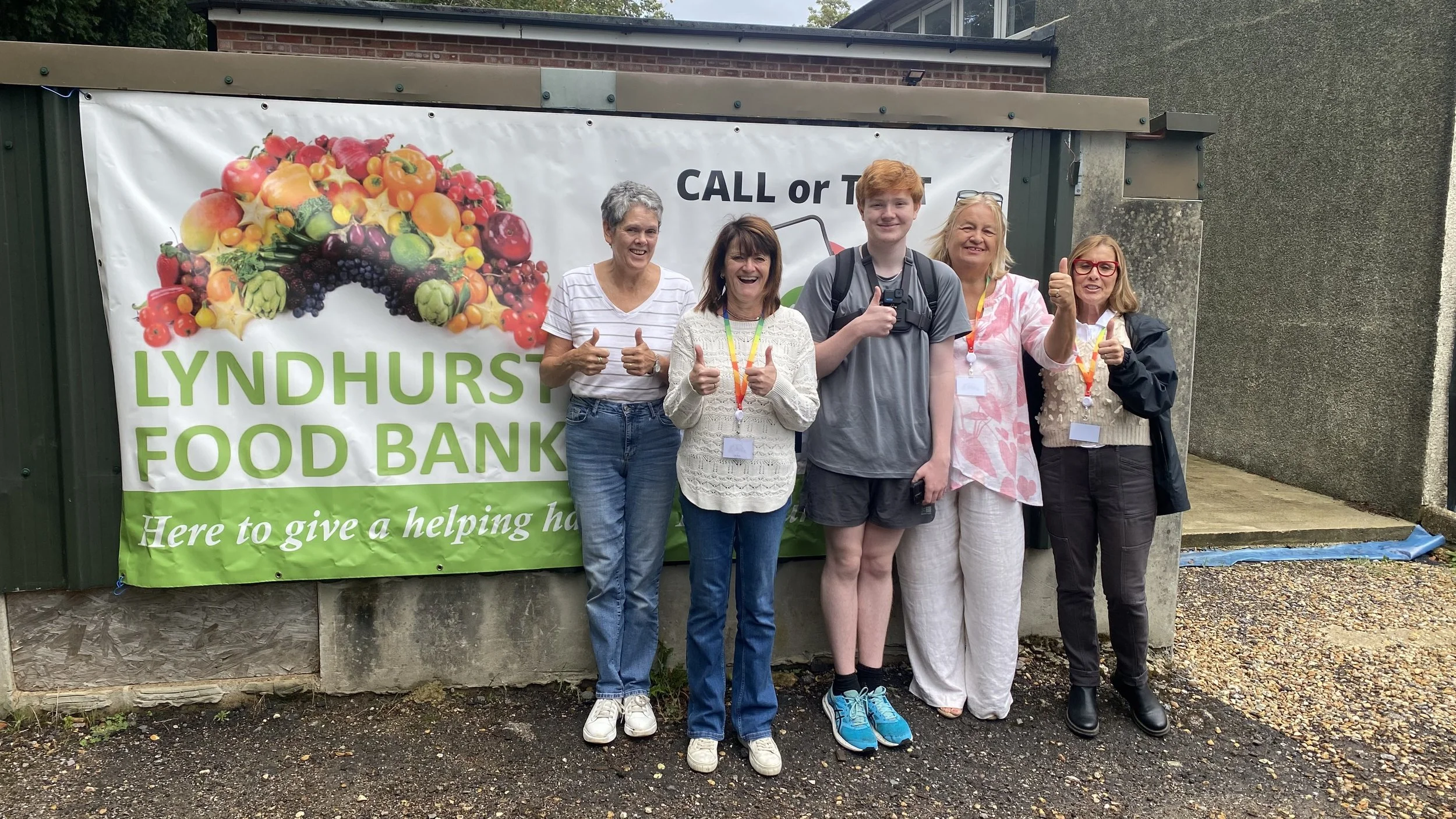 Group of five people standing in front of a banner for Lyndhurst Food Bank, smiling and giving thumbs up.
