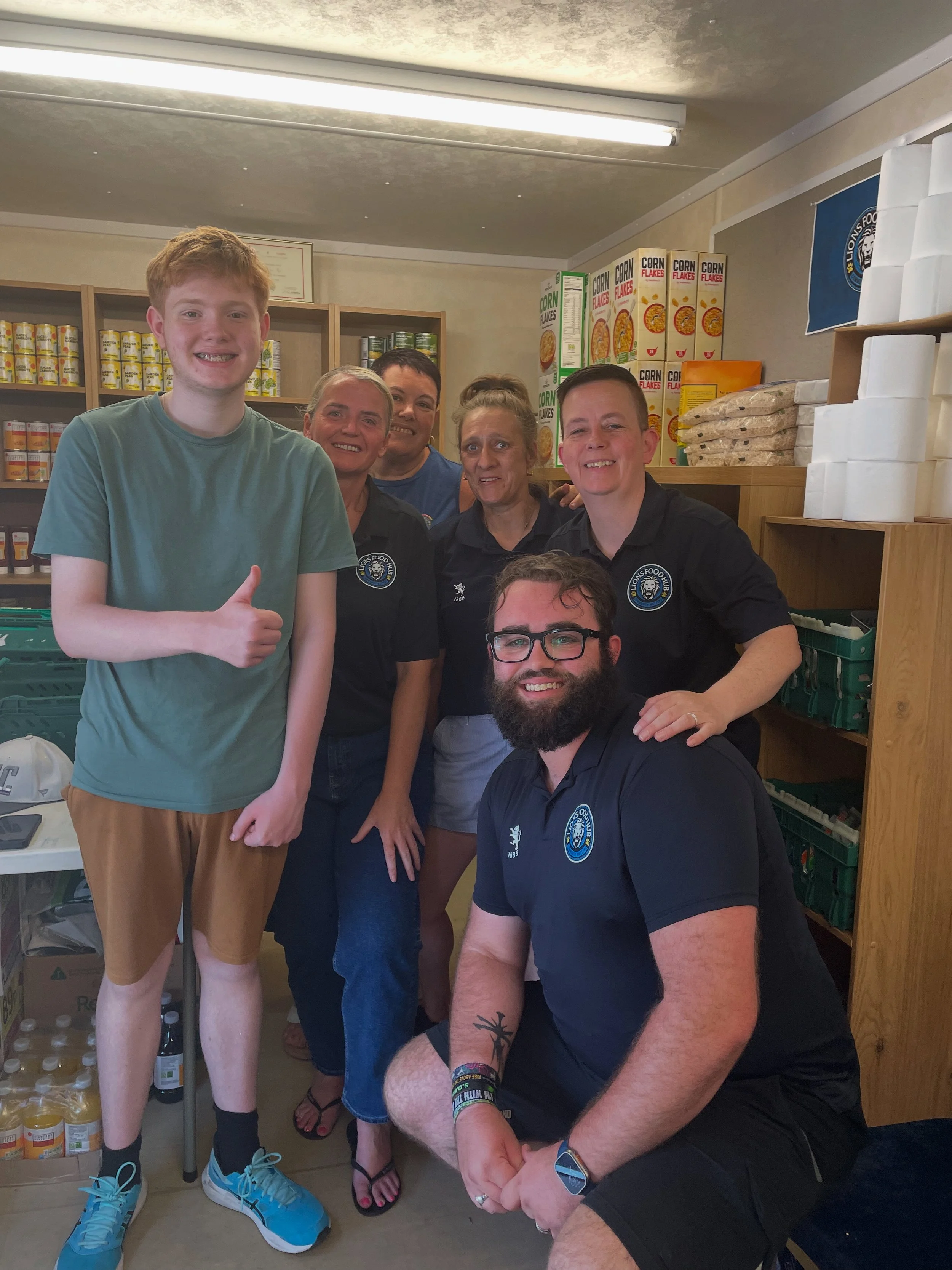 A group of six people smiling and posing together in a room with shelves filled with canned and packaged food. One person in the front is giving a thumbs-up and wearing sneakers, while the others are standing or crouching behind.