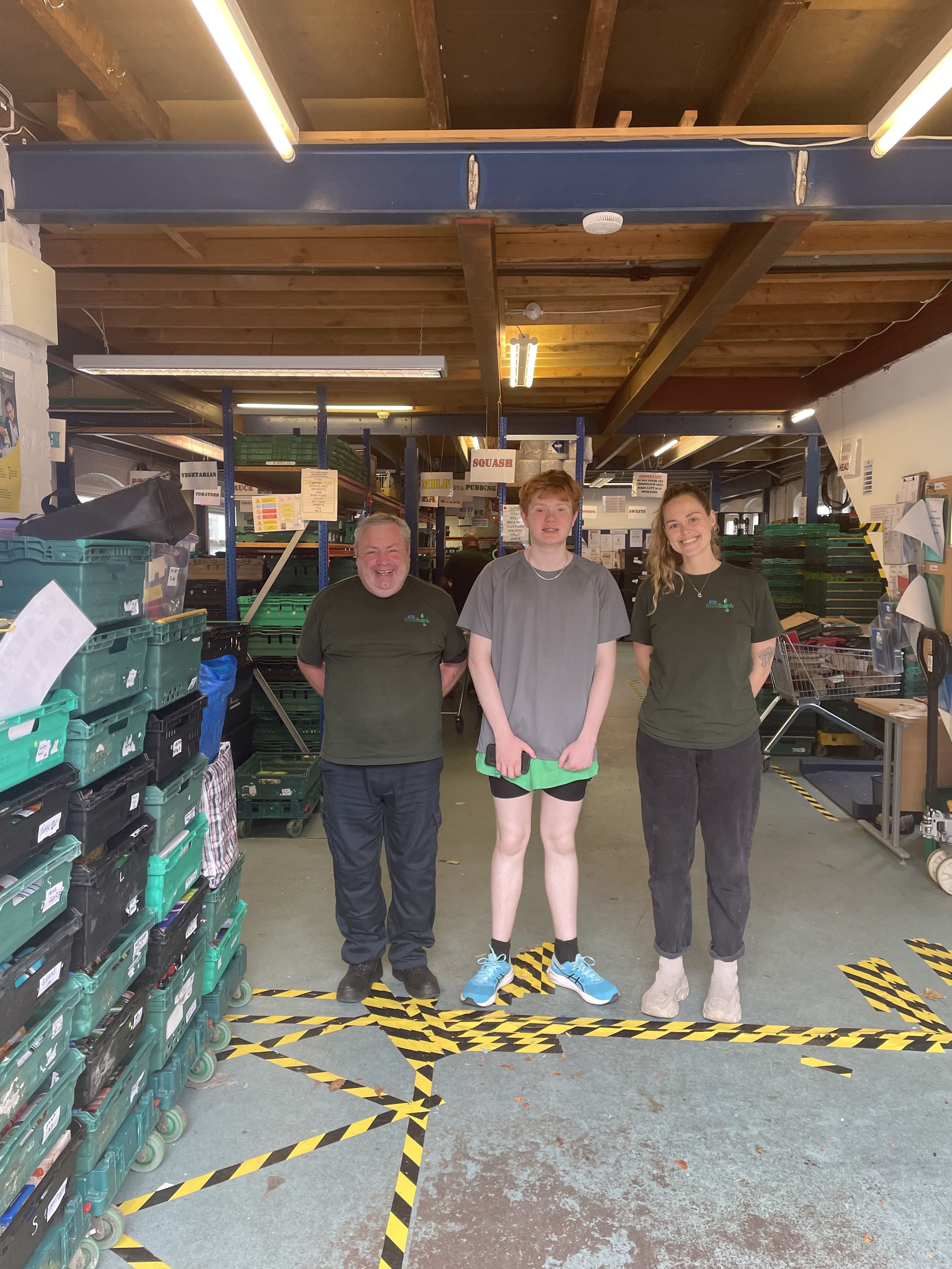 Three people standing in a warehouse, smiling at the camera. The warehouse has shelves with labeled bins, and the floor has yellow and black caution tape markings.