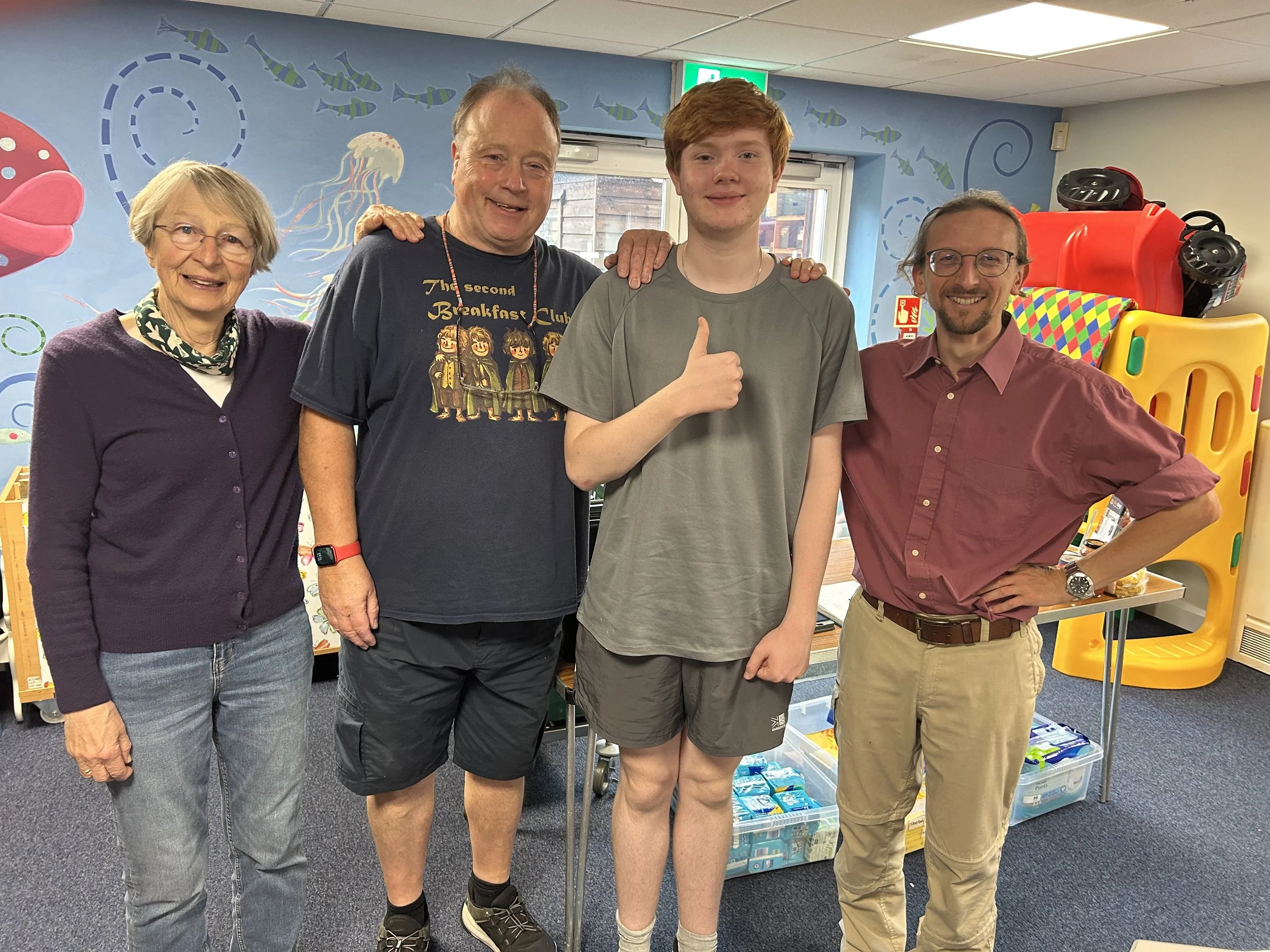 Five people standing together indoors in front of a colorful aquatic-themed wall. The group includes a woman with glasses and short gray hair, a man wearing a T-shirt, a young man with red hair, another man with glasses, and another man with glasses and a maroon shirt. Behind them are large plastic toys and storage bins.