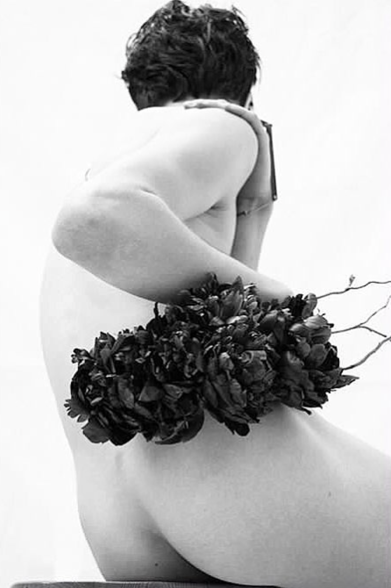 A black and white photo of a person with short hair, resting their head on their arm. The person is sitting with their back partially exposed, with a floral accessory covering their lower back.