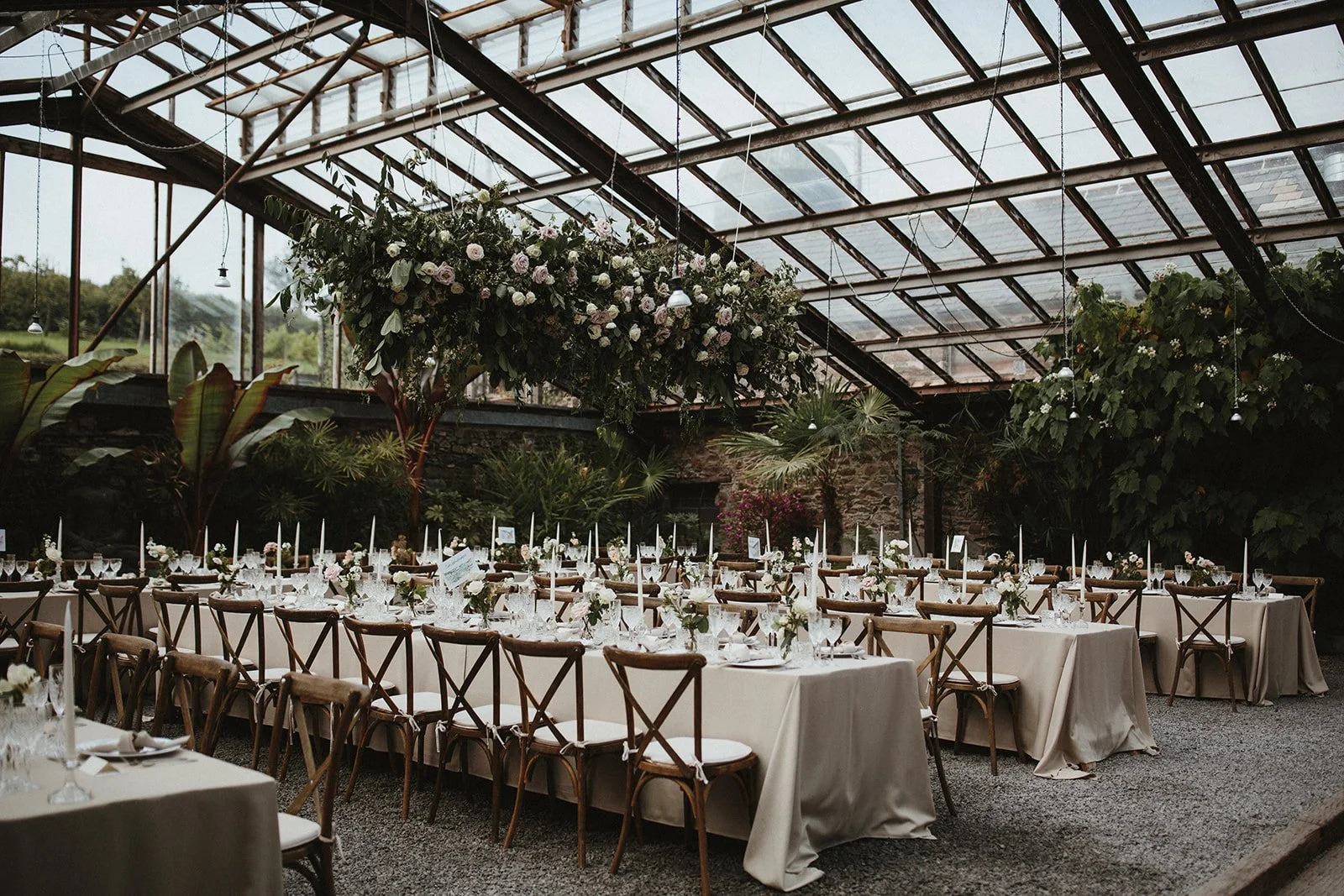 Long banquet tables with white tablecloths and floral centerpieces arranged in a greenhouse with large windows and glass ceiling. Wooden chairs are lined up along the tables, and the setting appears to be prepared for a formal event or celebration.