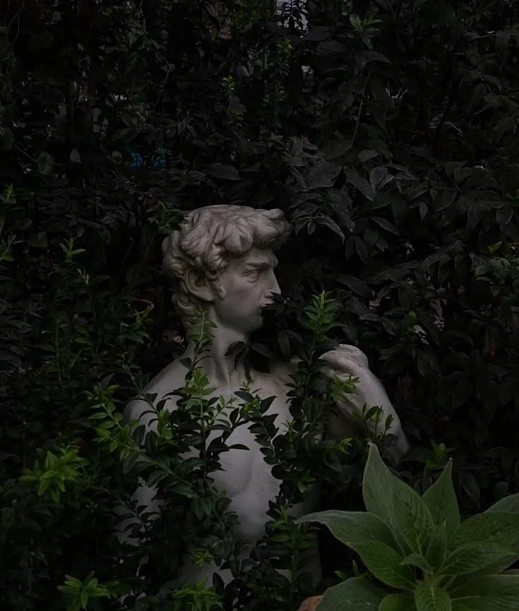 A classical marble bust of a man partially covered by dense green and dark foliage.
