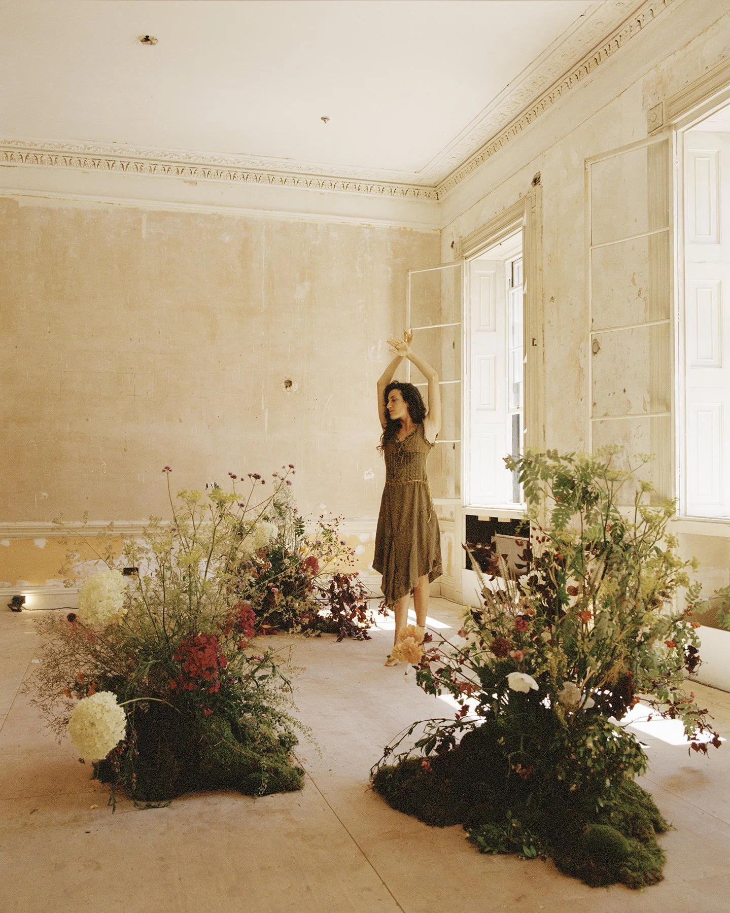 A woman in a brown dress standing in a decorated room with large floral arrangements in the foreground, and windows with white shutters in the background.