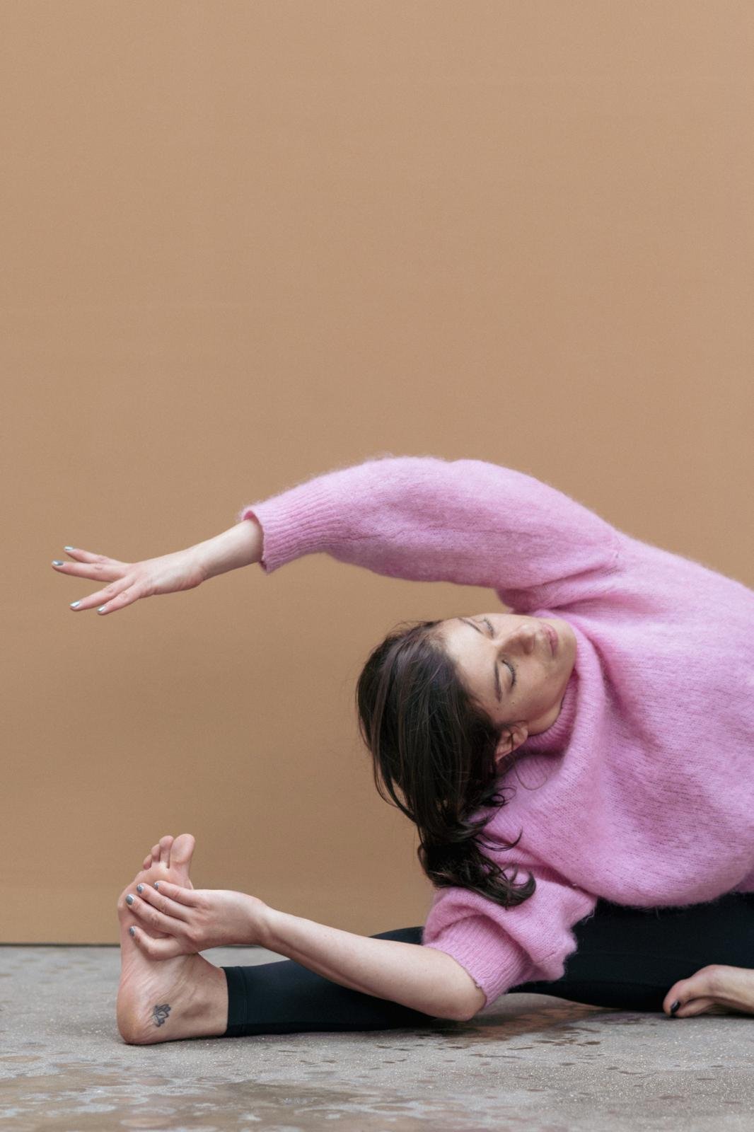 Femme faisant une pose de yoga sur un tapis au sol, portant un pull rose, contre un mur beige, avec les yeux fermés.
