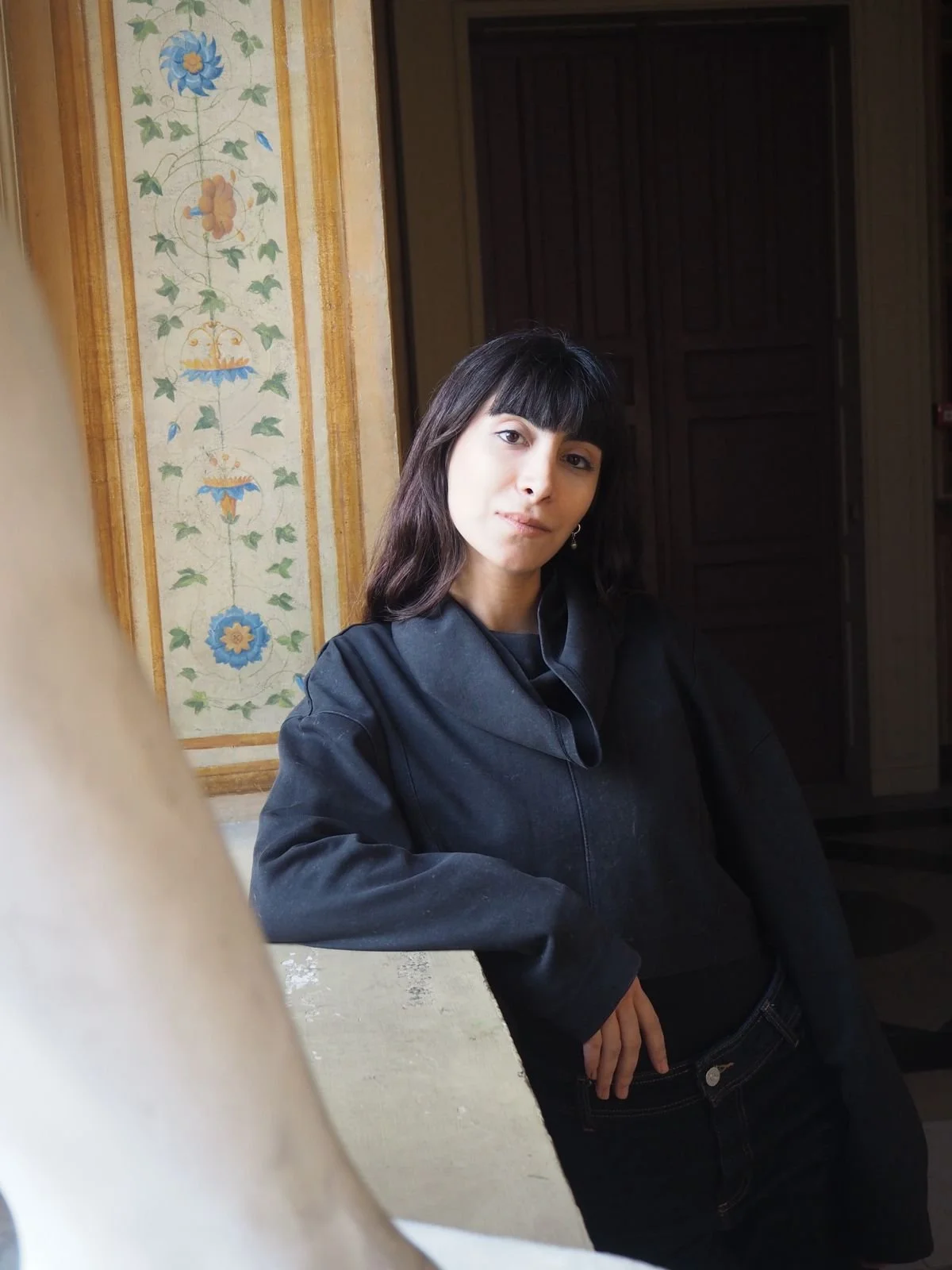 Une femme aux cheveux longs et noirs pose devant un mur décoré avec des motifs floraux, porte un vêtement noir, et regarde vers l'objectif.