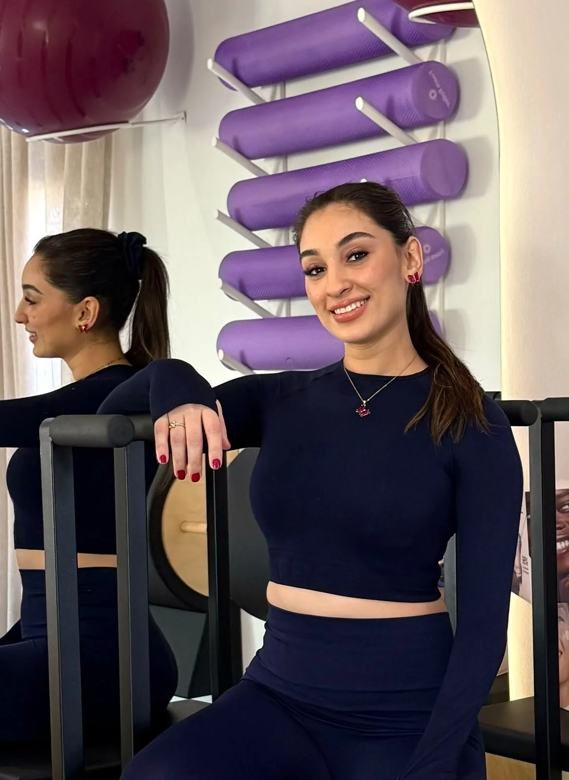 Une jeune femme souriante avec un pantalon et un haut noir, avec des écouteurs en forme de cœur rose, pose dans une salle de sport ou de fitness, avec un mur à ballons violets en arrière-plan.