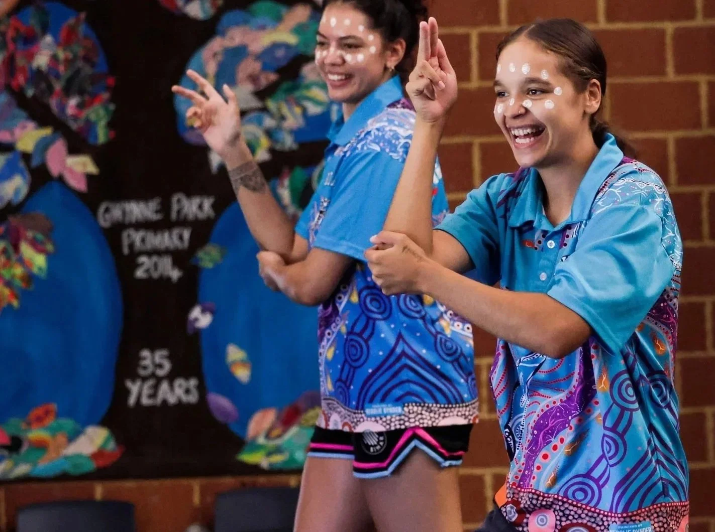 Two young women in colorful blue shirts smiling and making gestures, standing in front of a decorated blackboard with a tree and the words 'Crime Pack' and '35 Years' written on it, in a room with brick walls.