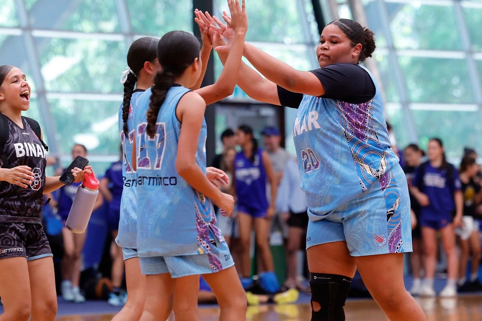 Young female volleyball players high-fiving during a game, with other players and spectators in the background.