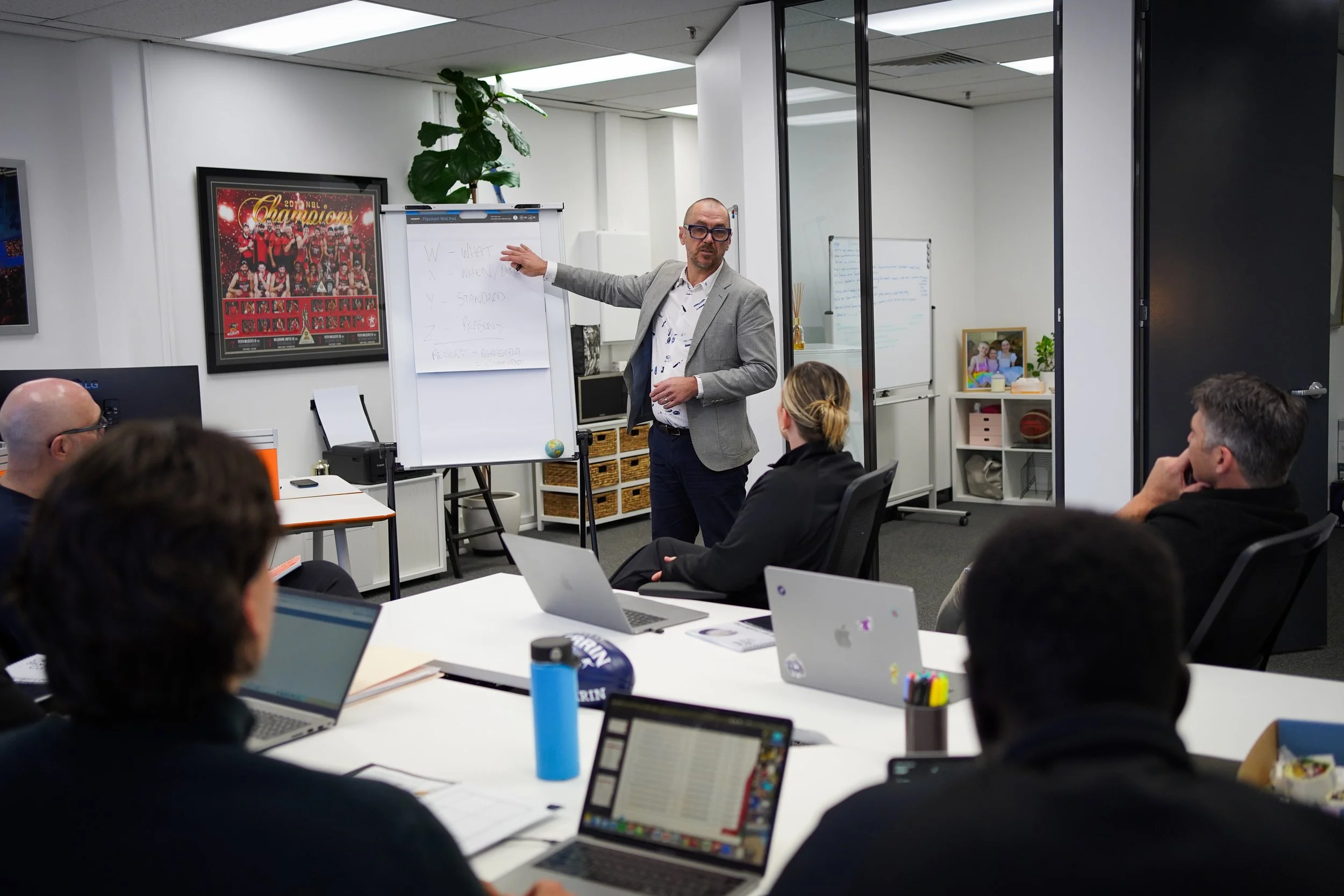 A man in a gray blazer giving a presentation to a group of people in a modern office conference room. The man is standing near a whiteboard with writing, while participants are seated at a table with laptops.