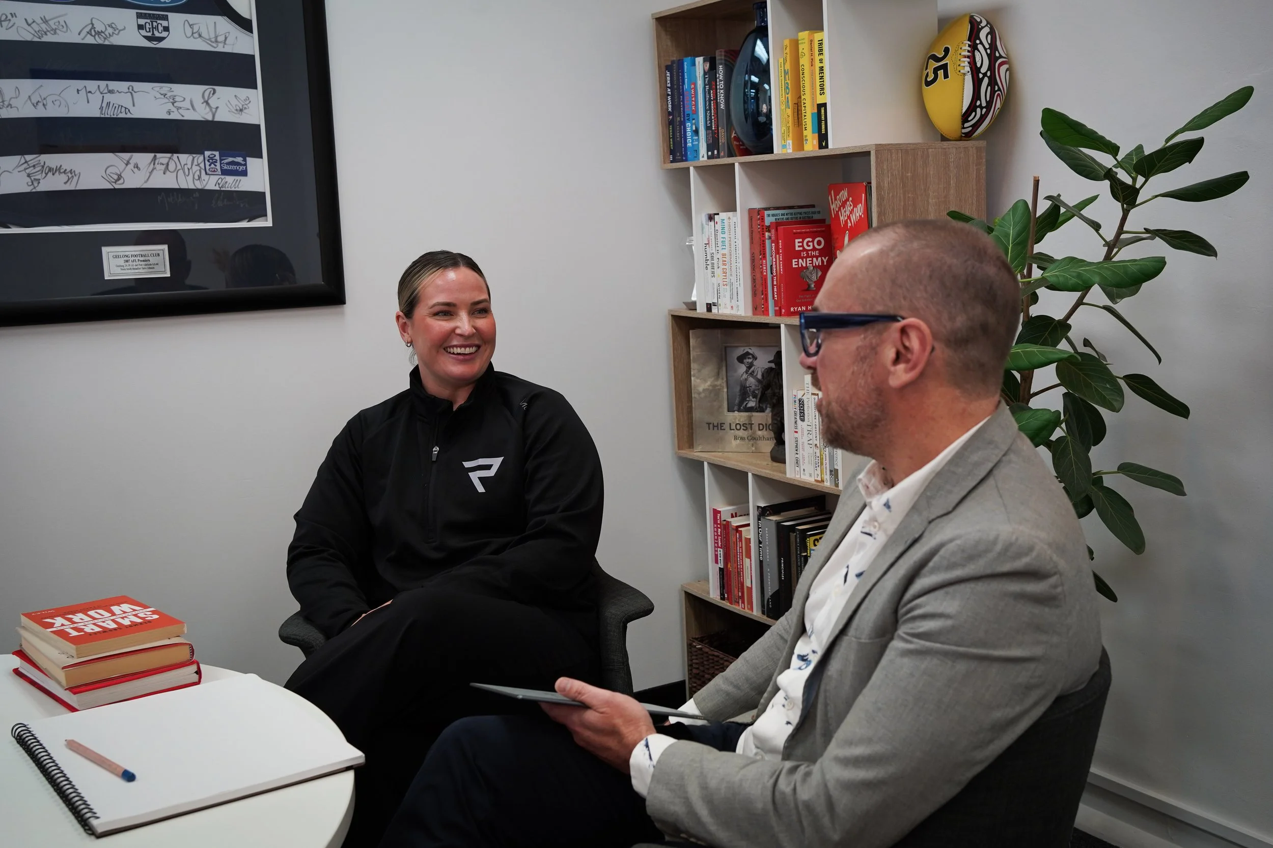 A woman with blonde hair in a black jacket smiling and talking to a man with glasses and a suit in an office setting. There is a bookshelf with books, a football, and a plant in the background. Books and notebooks are on the table.