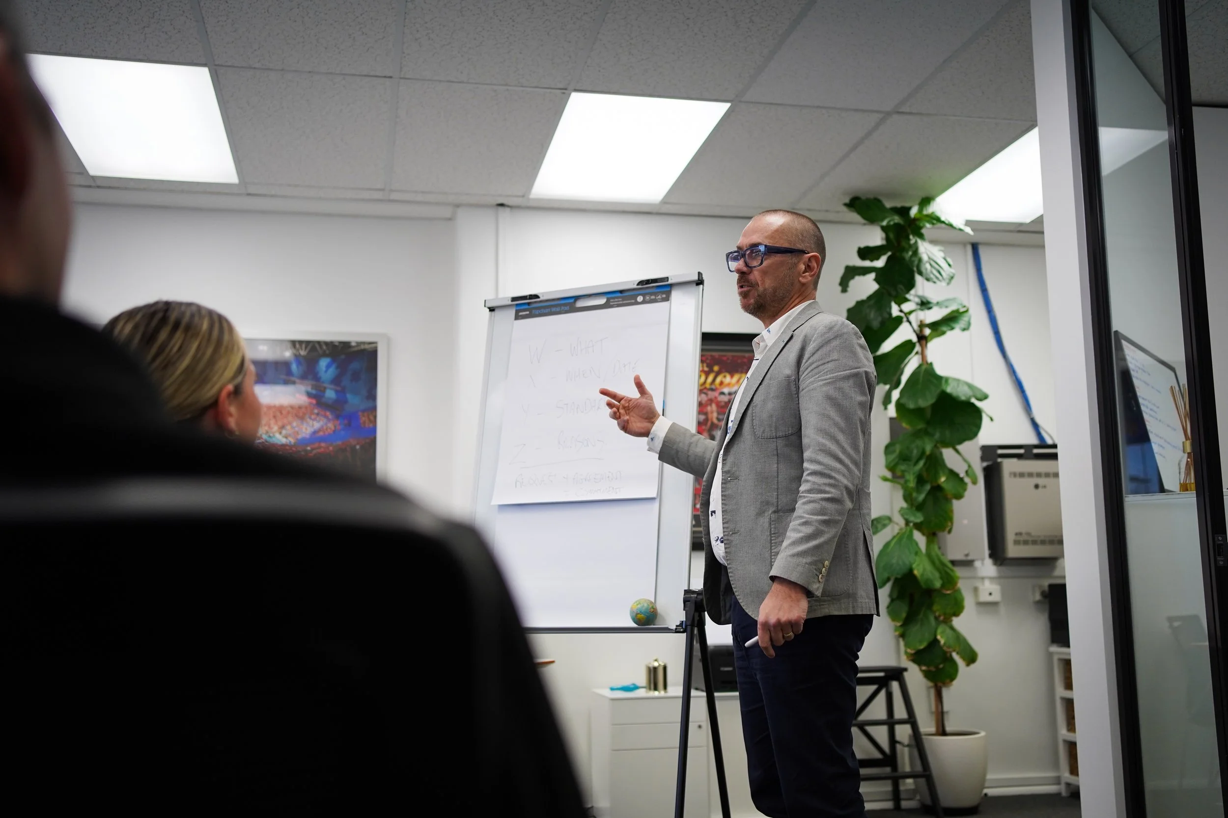A man with glasses, wearing a gray blazer, giving a presentation in an office classroom. He is standing next to a whiteboard with writing, and there is a large plant behind him. Several people are seated, watching him.
