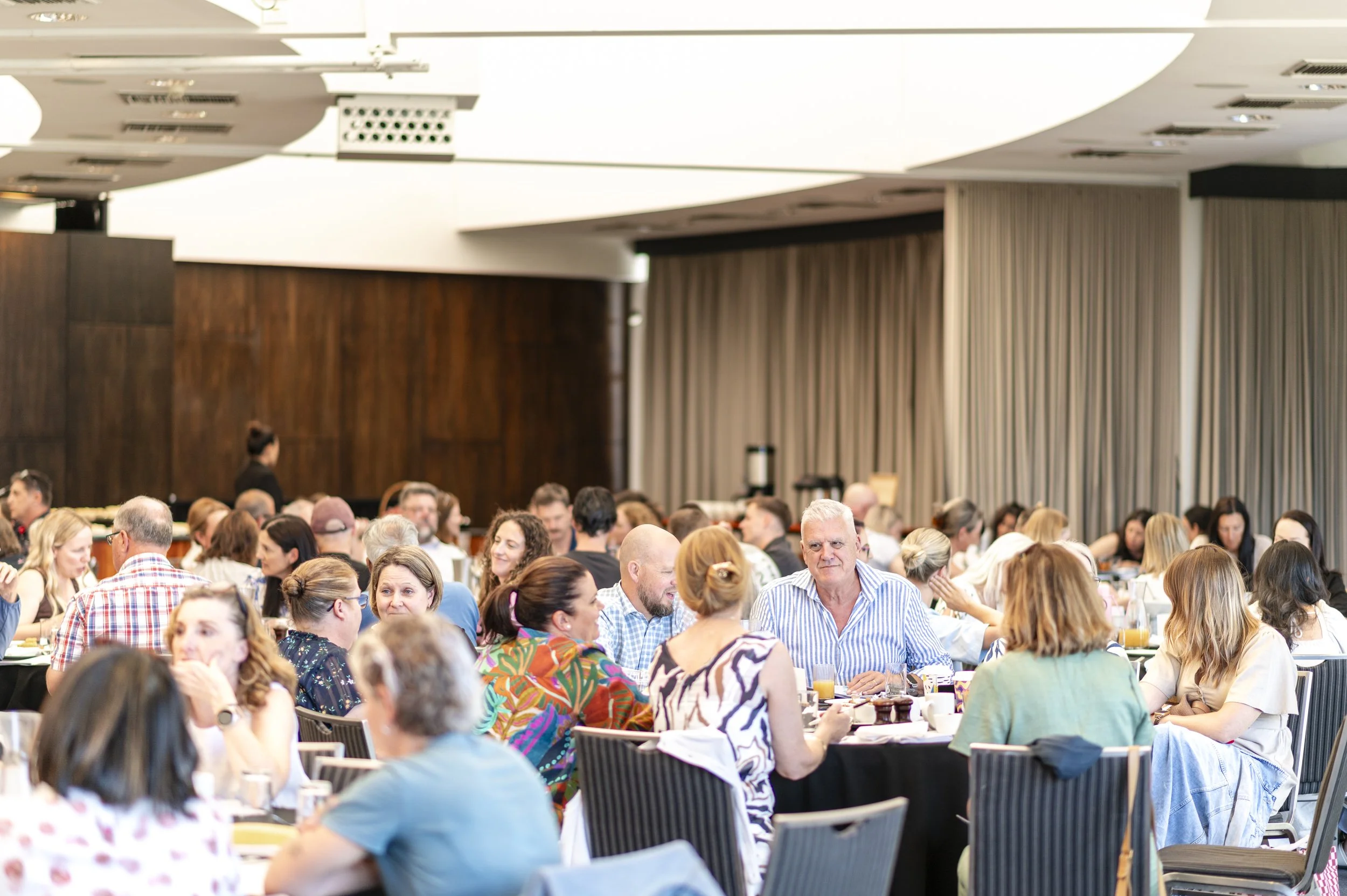 A large group of people attending a conference or meeting in a spacious, well-lit room with modern decor, wooden panels, and vertical blinds.