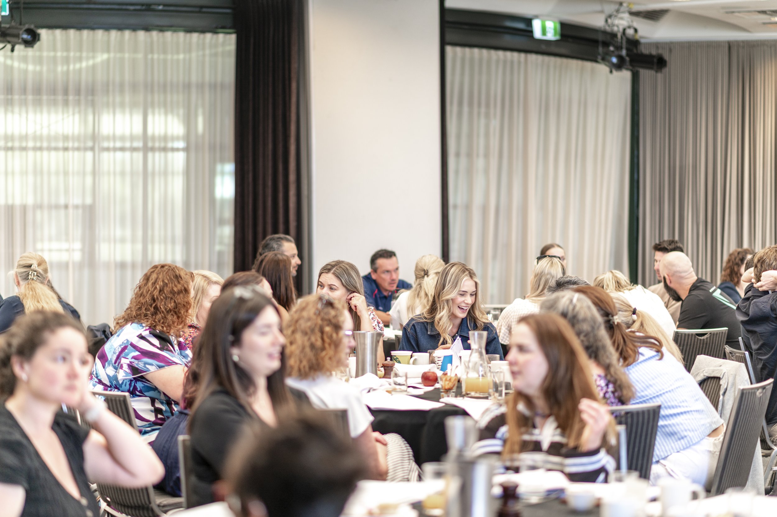A large group of people sitting at tables in a conference or meeting room, engaged in conversation and smiling, with beige curtains and large windows in the background.