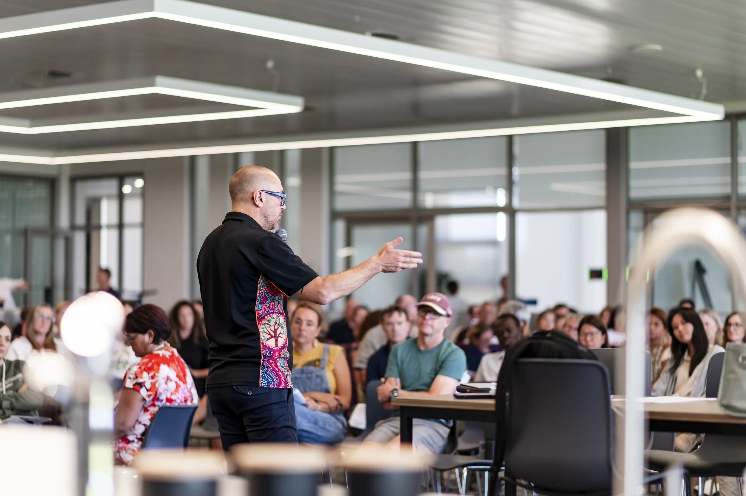 A man with glasses and a black shirt with a colorful pattern on the side is speaking into a microphone in front of a large audience in a modern conference room with glass walls and ceiling lights.